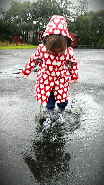 Chloe in her bright red rain coat splashing in puddles on a very grey, wet day in Manchester