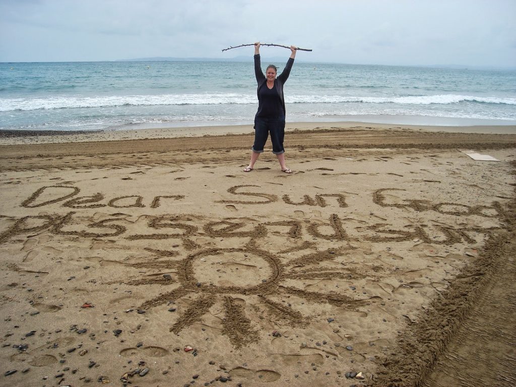 Should term time holidays be allowed?
Colette standing on the beach in Spain on a very grey, cloudy day in front of some writing on the sand. The writing says, "Dear Sun God, Pls send sun"