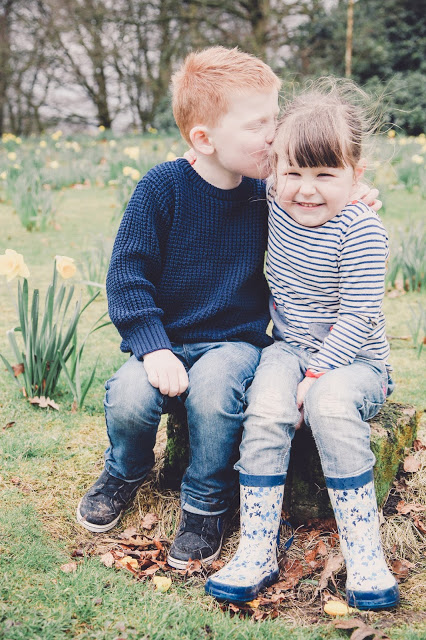 Brother and sister sitting on a tree stump in Heaton Park. Big brother is kissing his little sister on the cheek. Photograph by Sally Langstaff