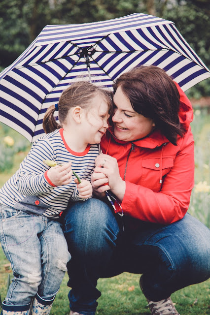 Mum wearing bright red rain coat and holding a navy stripe umbrella with daughter