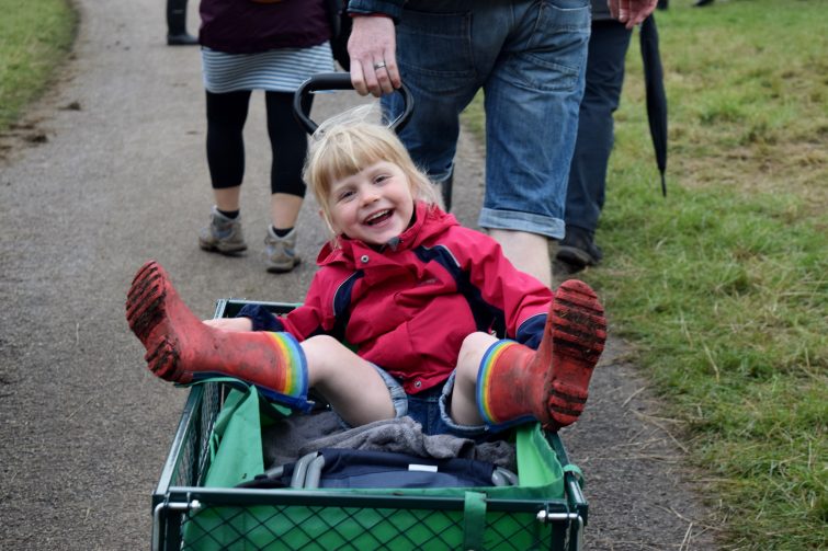 Little girl sitting in a festival trolley wearing brightly coloured wellies and w waterproof coat