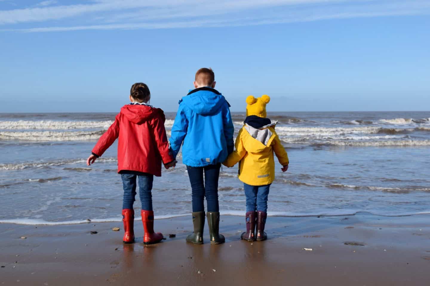 Three children wearing bright coats and paddling in the sea at National Trust Formby