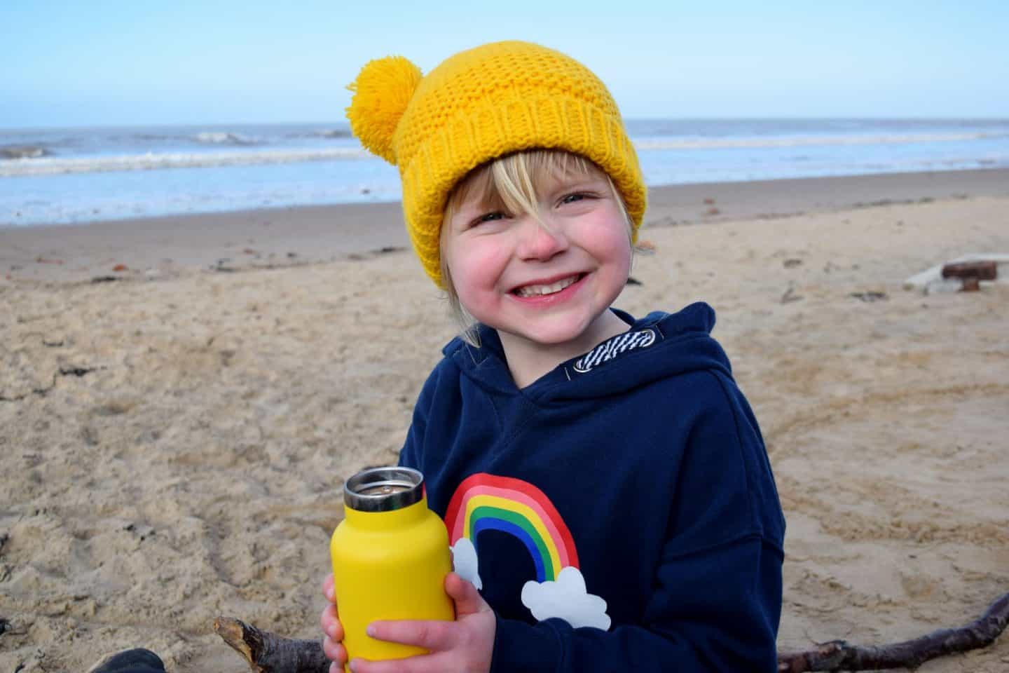 Drinking hot chocolate on the beach at Formby wearing a bright yellow hat and rainbow jumper