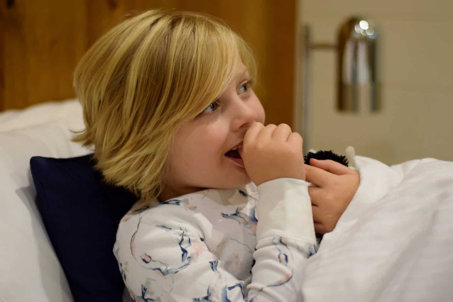 Little girl tucked up in bed, sucking her thumb and watching a film with her cow teddy