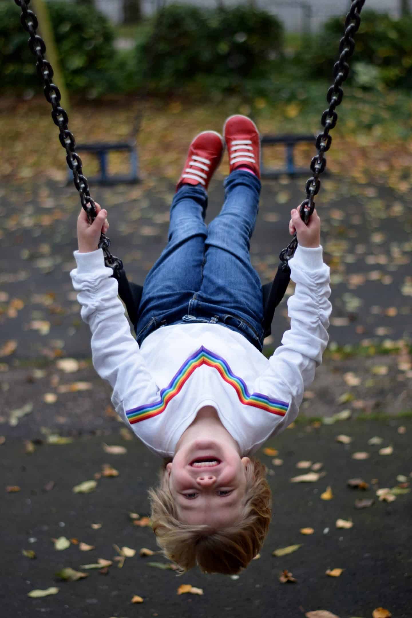 Little girl hanging upside down on a swing wearing a rainbow sweatshirt and red trainers