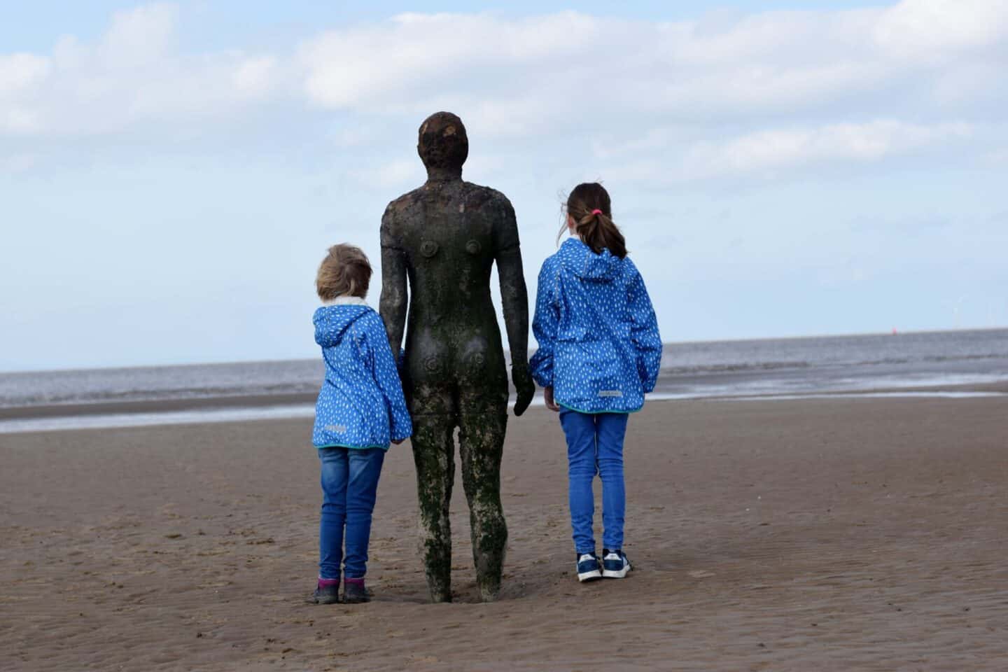 Girls wearing Muddy Puddles coats and standing next to Another Place statue
