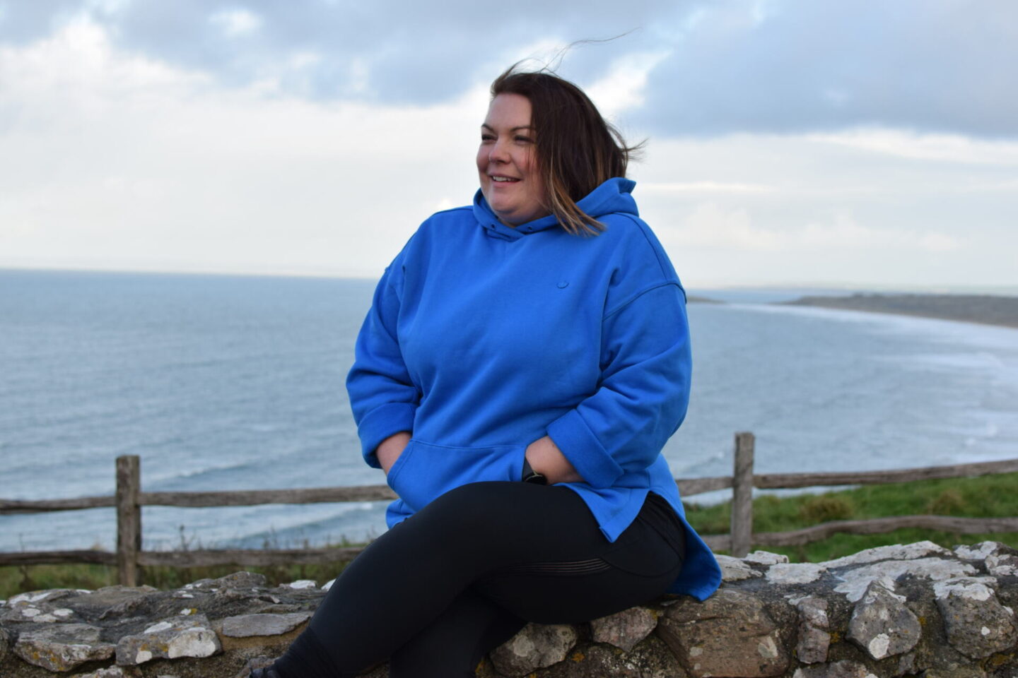 Colette wearing Adidas hoodie, sitting on a wall at the coast and looking windswept!