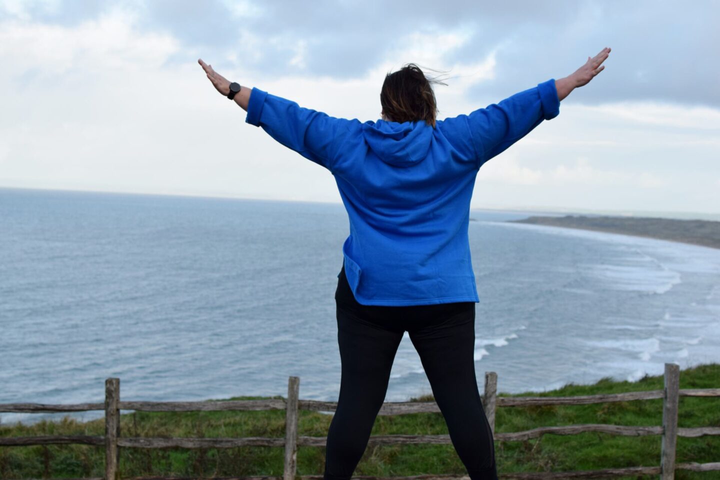 Colette standing on a wall facing out to sea with her arms and legs spread.