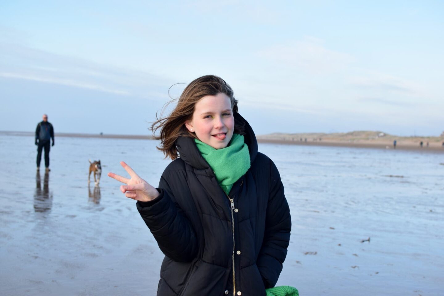 Chloe standing on the beach doing her standard tongue out, peace sign pose wearing a black duvet coat and cosy green scarf. Dad is looking on in the background but out of focus.
Chloe will be changing secondary school in Year 7