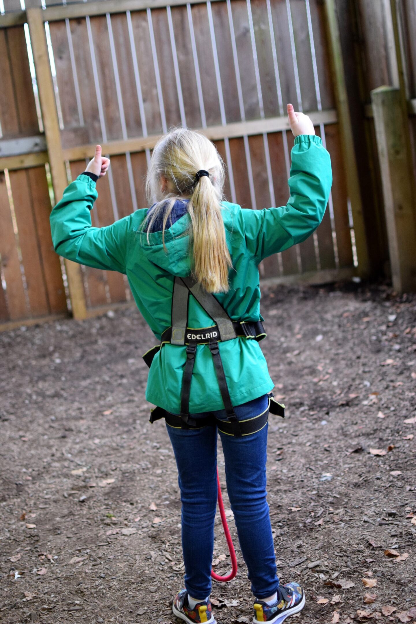 Amy has her thumbs in the air and her back to the camera demonstrating that her safety harness fits properly and she's ready to get up in the trees