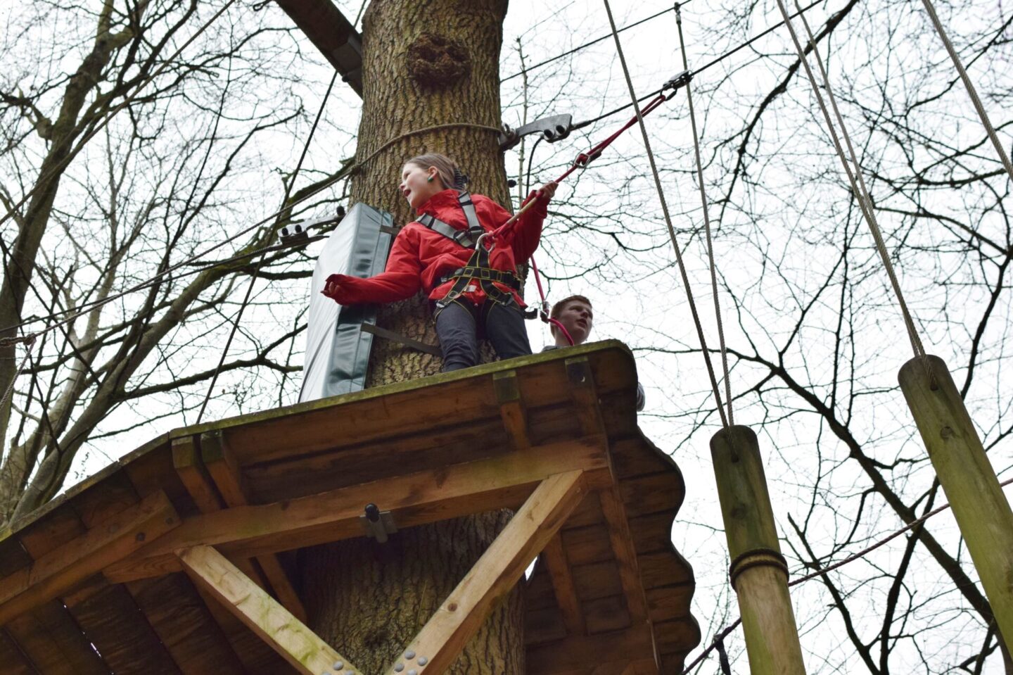 Chloe stands on a high wooden platform waiting her turn to do the next aerial obstacle, shouting words of encouragement to her little sister.