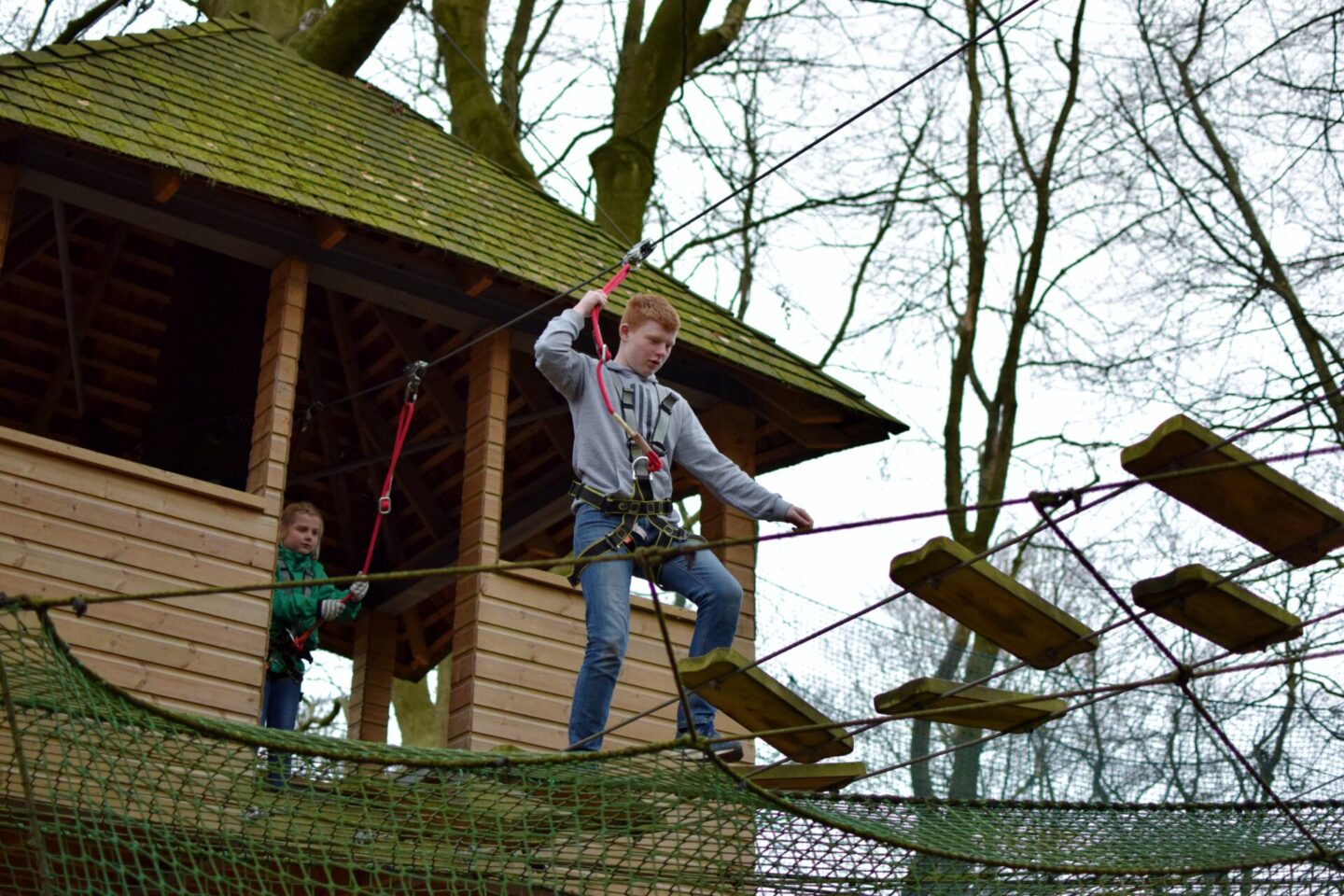 Ben is stepping confidently across a wooden aerial obstacle at TreeTop Manchester whilst his little sister watches on.