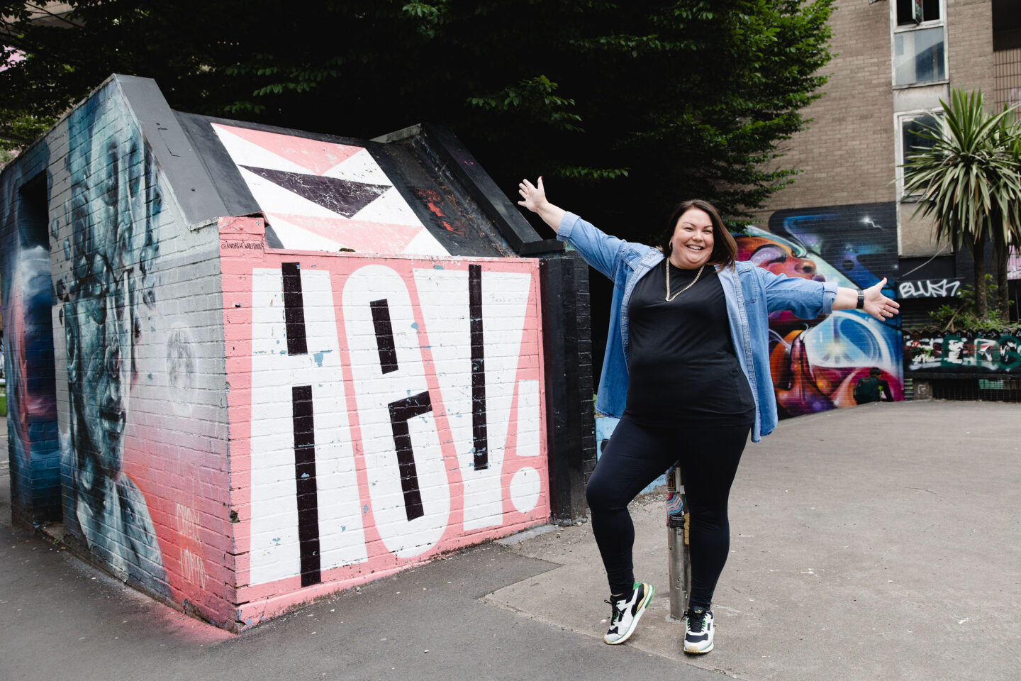 Colette standing in front of street art in the Northen Quarter - the street art reads HEY and Colette is standing with her arms wide open and a big cheesy grin