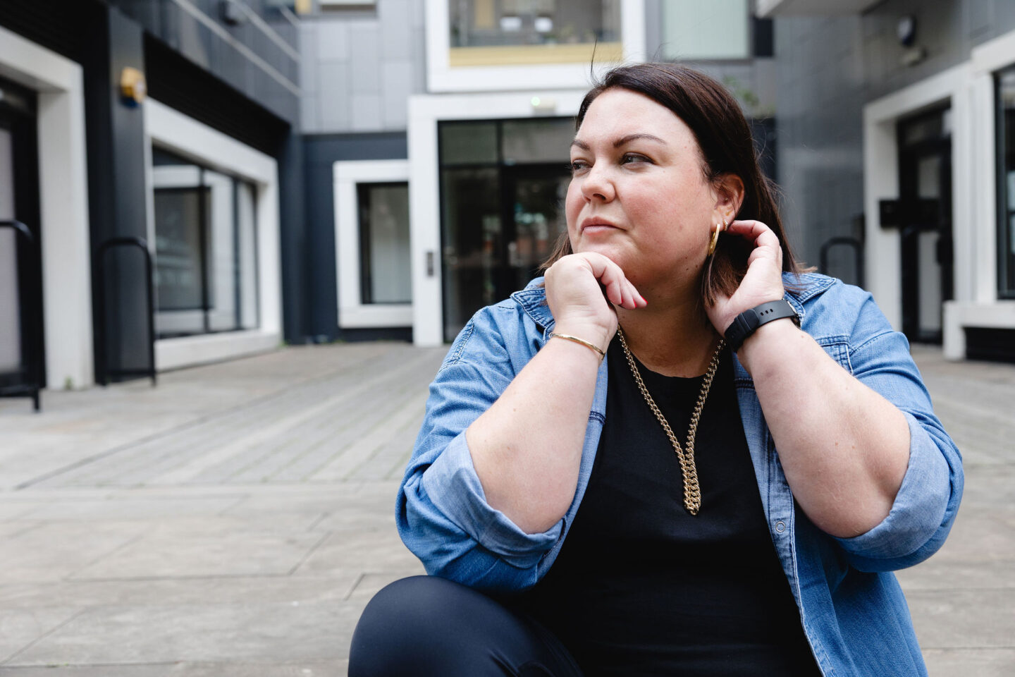 Colette B wearing a denim shirt rolled up at the sleeves, resting her chin on her hand and looking thoughtful.